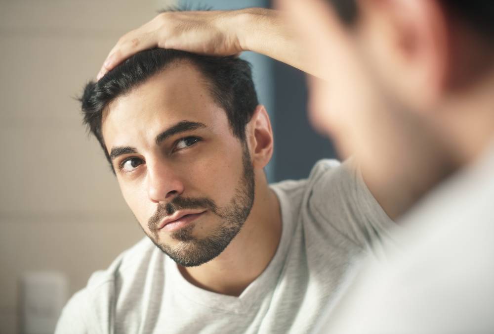 Man looks into bathroom mirror to examine his receding hairline while considering non-invasive treatment options for hair restoration.